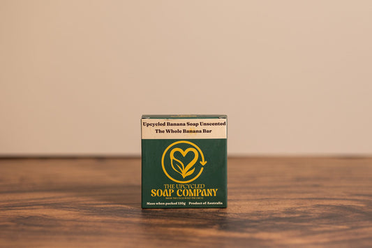 A single packaged soap bar standing upright on a wooden surface against a plain beige wall. The package is dark green with a large gold leaf-and-heart logo and white text reading 'The Unplugged Soap Company' and 'Upcycled Banana Soap Unscented The Whole Banana Bar.