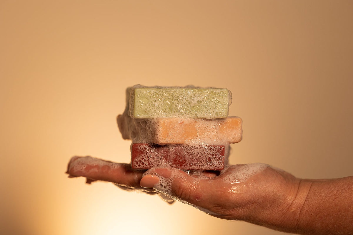 A hand holding a small stack of four frosted soap bars in different colors—light green, peach, dark red, and terracotta—against a warm beige background.